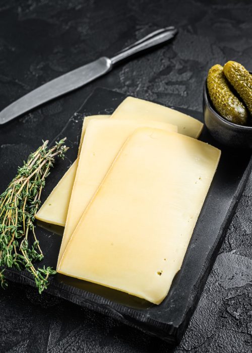 Fresh Uncooked Raclette Swiss cheese slices on marble board. Black background. Top view